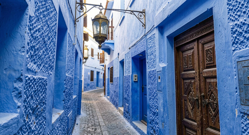 A narrow blue painted alley in a Moroccan medina with a hanging brass lantern and a carved wooden door.